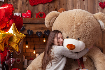 attractive young woman in a beautiful background for a photo shoot on Valentine's Day, cuddling with a huge teddy bear, a wooden fireplace with candles and flowers, and tree with red and pink hearts.