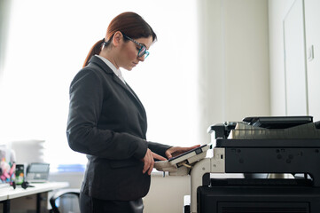 Business woman in a suit and glasses makes copies of documents on a photocopier. Female office manager is doing paper work. Secretary makes photocopies.