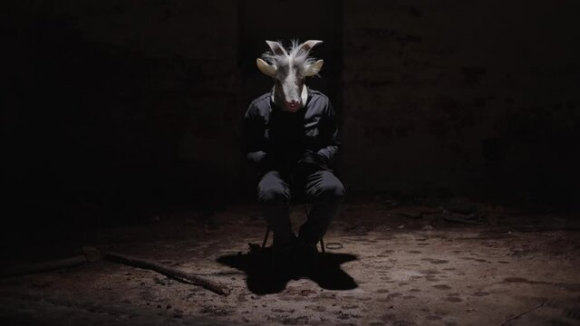 Creepy masked person sitting in an abandoned basement. Scary scene of dangerous stranger looking at the camera. Haunted house with a masked maniac inside.