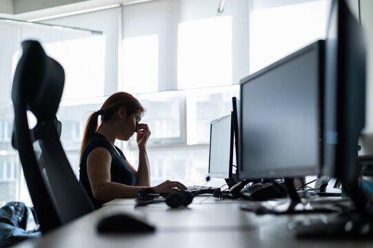 Tired woman workaholic t-shirt works in an empty office and holds fingers on the bridge of the nose. A girl with a headache works overtime. Female silhouette on a window background. Problems at work.