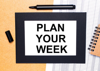 On a wooden table, there is a black open marker, a brown notepad and a sheet of paper in a black frame with the text PLAN YOUR WEEK. View from above.