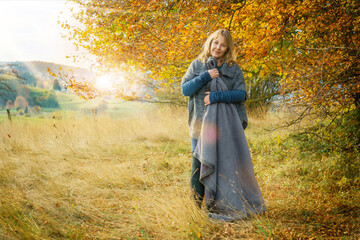  A woman of 40 years in autumn nature landscape. The blonde woman wears clothes made of wool and boots. She has a blanket. - obrazy, fototapety, plakaty