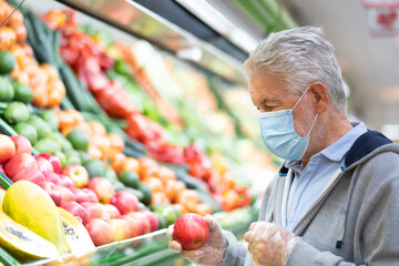 Senior adult man with surgical mask due to coronavirus while shopping at supermarket. White-haired retiree choosing apple-fruit  in the vegetable and fruit department. Consumerism concept