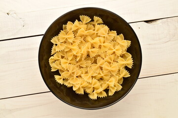 Light yellow pasta, in the form of a butterfly in a black ceramic plate on a white wooden table, close-up, top view.