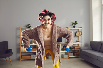 Happy young woman enjoying sports workout at home during lockdown. Funny crazy housewife in homewear, hair curlers and beauty skincare face mask doing fitness exercise with dumbbells in living-room