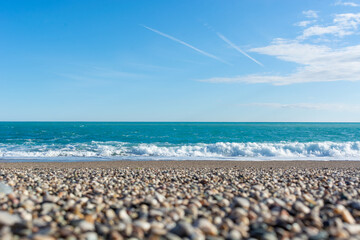 Pebble beach from a lower angle and a blue sea with waves in Antalya under a blue sky with white clouds and plane tracks in sunny weather. No people. Close up. Blurry foreground