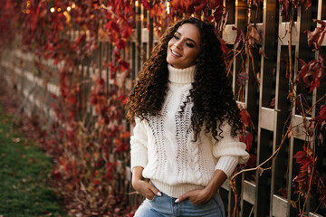 Against the background of the wall of wooden beams entwined with red ivy stands flirtatiously smiling curly brunette in a white sweater sticking hands in the front pockets of jeans. High quality photo
