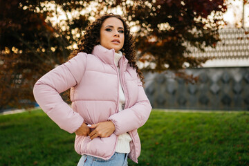 Obraz premium A European girl with dark curls in a pink jacket and jeans walks in the autumn park on the green grass. High quality photo