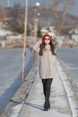 A girl in a gray coat, red hat and black jeans poses on the pier. Winter. Coldly. Ice. Sunny. 