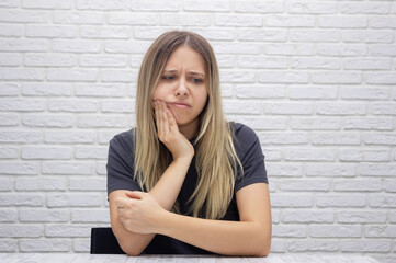 A young beautiful blonde woman with a toothache is holding her cheek against the background of a white brick wall. The girl suffers from a toothache, waiting for a doctor's appointment and is worried