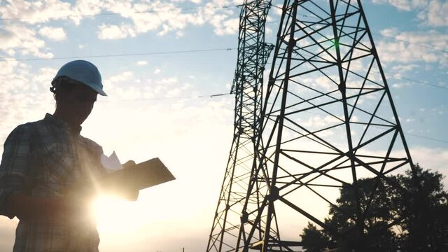 silhouette of an electrician engineer working next to an electrical support tower energy. electricity high voltage tower support. electrical engineer sunset inspect voltage electricity pylon