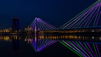 Backlit bridge at night and blurred reflected in the water. Vinogradovsky cable-stayed bridge over Yenisei river in Krasnoyarsk, Russia.