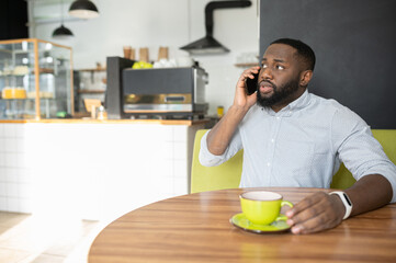 Fototapeta na wymiar Puzzled African-American guy is sitting in the cafe with cup of coffee and has serious phone conversation, a multiracial man talking on the smartphone