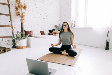 Beautiful brunette fitness woman meditate in front laptop, doing yoga indoors at home. Staying fit and healthy