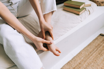 Obraz na płótnie Canvas Woman sitting on the bed massages her foot, close-up. Woman with a slender body massages the leg, Young woman massaging her foot on the white bed after training or hard working day. Healthcare concept
