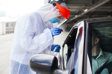 Medical worker in PPE performing nasal & throat swab on person in vehicle through car window