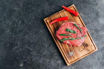 Raw beef steak on wooden board and black background with rosemary, chili pepper and spices, copy space.