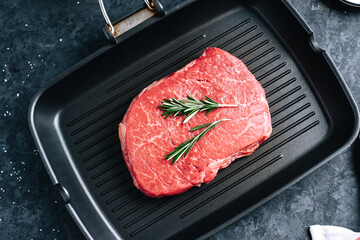 Raw beef steak on black grill pan with rosemary, top view.