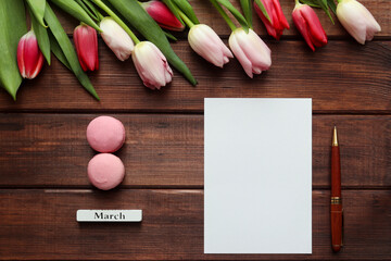 Women's Day template for greetings. Beautiful bouquet of tulips on a dark wooden background with empty blank of paper, pen and eight-shaped macarons. Copy space for text. Top view, flat lay