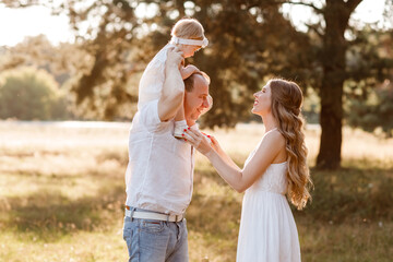 Portrait of a happy young family spending time together in the summer nature, on vacation, outdoors. Mom, dad holds daughter on shoulders stand on the grass. The concept of family holiday.