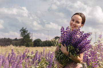 Beautiful smiling girl with a bouquet of lupine flowers. A girl with a bouquet of lupins stands on a flower field. Young brunette girl. Summer time. Lupine field