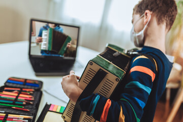 Focused boy playing accordion guitar and watching online course on laptop while practicing at home....