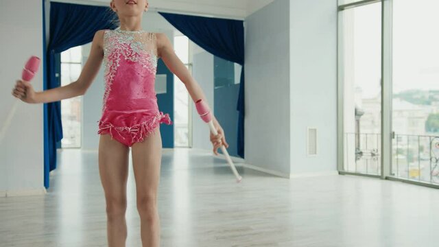 Little girl in performs exercises with clubs for rhythmic gymnastics and makes wheel catching object in air.