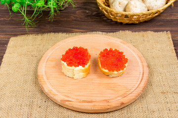 Delicious sandwiches with red caviar and butter on a wooden board against a background of herbs and mushrooms.