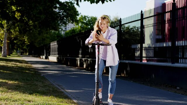 Beautiful blonde hair lady with the red headphones in the middle of the street take a stop with her electric scooter to texting someone. 4k
