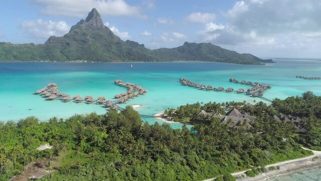 Aerial - Panoramic view of luxury overwater villa resort in Bora Bora with a view toward Mount Otemanu. Tilt down toward bungalows