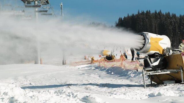 Snow making system. Snow cannon makes artificial snow of water on sunny day in ski resort. On backdrop skiers slide down snow-covered white slope of mountain, ski lift working, winter forest, blue sky