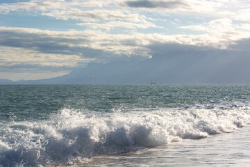 Raging waves on the sea beach and a ship in the distance with mountain views under blue sky with white clouds and sunbeams in Antalya in winter