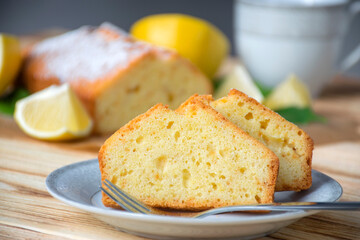 Piece of lemon cake on plate on rustic wooden board with full pie, lemons and cup on background. Easy recipe of citrus dessert for everyday cooking. Homemade bakery by classic recipe.