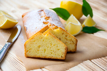 Loaf of traditional lemon pound cake with pieces of lemon, green leaves and knife on rustic wooden background. Slice of citrus pie by classic recipe. Homemade bakery, vegan dessert.