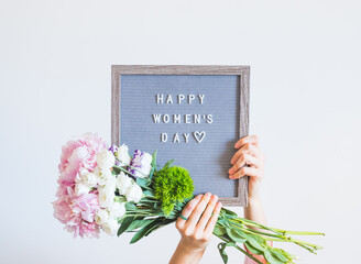 Young woman holding pink bouquet and letter board saying happy womens day on gray wall background
