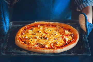 A delicious pizza with cheese, vegetables and mushrooms lies on a tray held by a chef in a blue apron and blue potholders. Home cooking.
