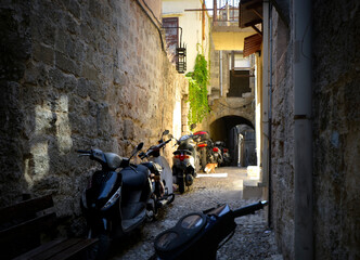 Fototapeta na wymiar A stray alley cat wanders down a narrow alley filled with motorcycles on the Mediterranean island of Rhodes Greece