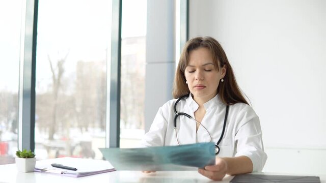 Young caucasian woman doctor wearing white medical coat and stethoscope holds the x-ray and makes notes
