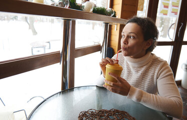 Young woman drinking from a straw and looking out the window at a snow covered nature while sitting at a wooden cafe