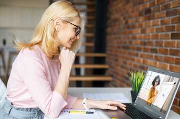 Intelligent mature blonde businesswoman in glasses sitting at the desk at home, studying, taking...
