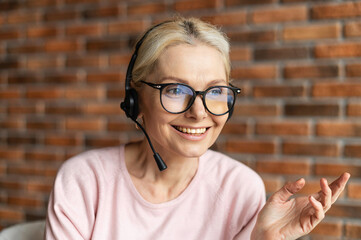 Close-up of the face of a charming beaming elegant mature blonde lady, wearing headset, glasses, smiling and looking away. Portrait of middle-aged customer service representative talking in the office