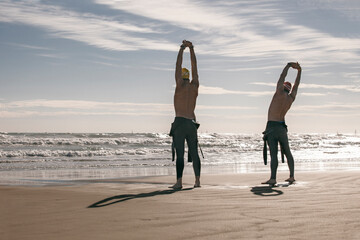 Two triathletes with wetsuit, cap and glasses stretching for swim on a sunny day,