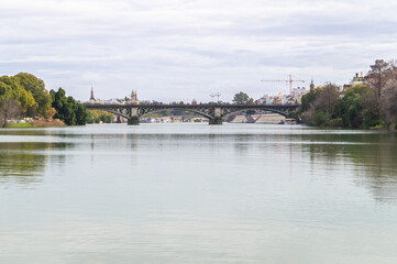 Urban landscape with the Guadalquivir river crossing the city of Seville in a cloudy day (Andalusia, Spain). Views of the Isabel II bridge, also known as the Triana bridge.