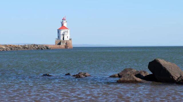 Lighthouse with red roof at the end of a pier on Lake Superior at Wisconsin Point. Large boulders surrounded by ripples of blue water being blown away from the camera by the wind. Slow motion clip.