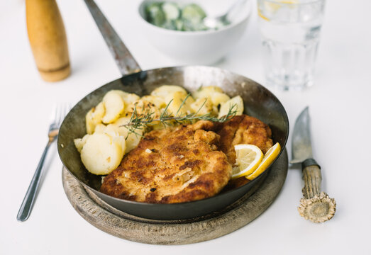 typical austrian wiener schnitzel with rosemary potatoes in iron pan and salad on white table