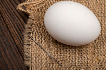 A chicken egg and a sewing needle on a homespun fabric with a rough texture.