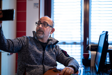 Middle-aged Caucasian man is sitting in his work station housed in the home kitchen. He takes a selfie. Work from home and technology concept.