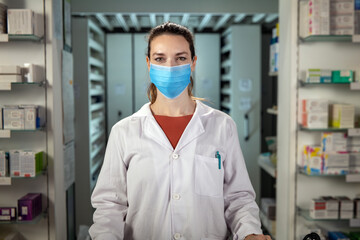 Fototapeta na wymiar Cinematic shot of female pharmacist consultant with protective medical mask is smiling satisfied with his job in camera in pharmacy. Concept of covid-19, protection, coronavirus, safety, health care.