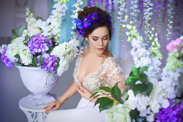 A beautiful young girl in a white lace boudoir dress, with blue anemones in her high hairstyle, sitting in a classic light bedroom decorated with hydrangeas, purple wisteria and other spring flowers.