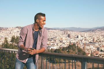 Black young man listening music with his phone and enjoying a nice panorama of Barcelona.